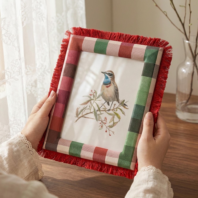 May include: A rectangular picture frame featuring a red and green checkered fabric border with a red fringe trim. The frame contains a print of a small bird with a blue throat perched on a branch with green leaves and small red berries.