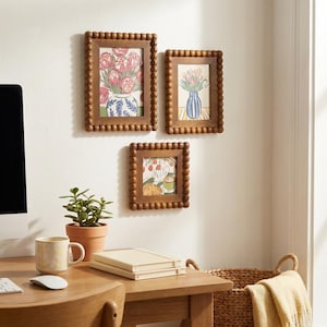 Può includere: Tre stampe artistiche incorniciate con cornici di legno a perline sono appese a una parete bianca. Le stampe raffigurano composizioni floreali in vasi. Una piccola pianta in vaso, una tazza e una pila di libri sono su una scrivania di legno in primo piano.