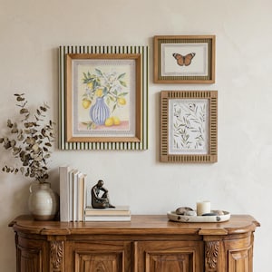 May include: Three framed art prints on a wall. The largest depicts lemons in a blue vase. The other two feature a butterfly and olive branch. The wooden frames have decorative borders. A wooden cabinet below holds books and a sculpture, with a vase and tray.