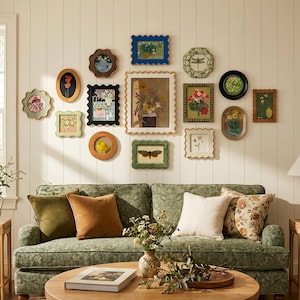 May include: A gallery wall featuring various framed art pieces with floral and nature-themed imagery. The frames are in various shapes and colors, including green, brown, and black. A green floral patterned sofa and pillows are in the foreground.