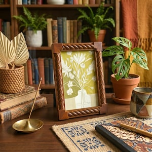 Może przedstawiać: Drewniana ramka na zdjęcia z obramowaniem w kształcie liny zawiera nadruk kwiatowy na żółtym tle. Ramka stoi na drewnianym biurku z tkaną matą, małą rośliną doniczkową i dekoracyjnym kubkiem. W tle znajduje się regał z książkami.