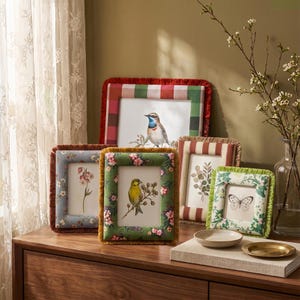 May include: A collection of five rectangular fabric-covered picture frames featuring various patterns including red and green plaid, floral prints, and stripes. Each frame is finished with a decorative fringe edge and displays botanical or bird art.