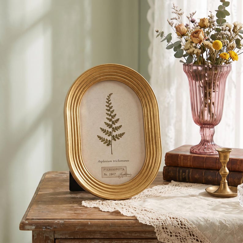 May include: An oval gold picture frame with a ribbed border containing a botanical print of a fern labeled Asplenium trichomanes and PTERIDOPHYTA No. 1847. The frame stands upright, displaying a detailed illustration of a green fern leaf on a cream background.