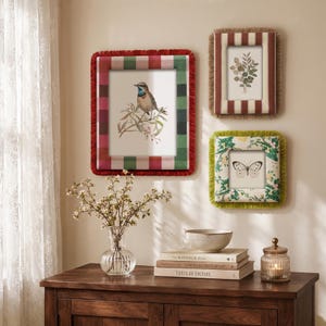 May include: Three rectangular wall frames with fabric borders and fringe. One large frame features a red and green plaid pattern with a bird illustration. Two smaller frames display a botanical print and a butterfly, with striped and floral fabric.
