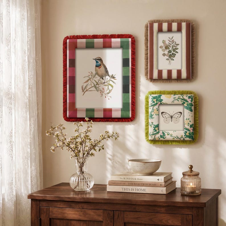 May include: Three rectangular wall frames with fabric borders and fringe. One large frame features a red and green plaid pattern with a bird illustration. Two smaller frames display a botanical print and a butterfly, with striped and floral fabric.
