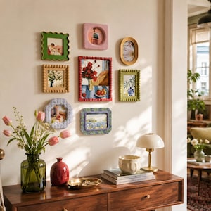 Puede incluir: Una composición de pared de galería con ocho marcos de fotos variados en diferentes formas, colores y texturas, incluyendo diseños festoneados, con cuentas y lisos. Los marcos muestran diversas obras de arte en una pared clara.