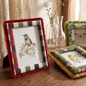 May include: A rectangular picture frame with a fabric border featuring a red, green, and white checkered pattern and red fringe trim. The frame holds an illustration of a bird with a blue throat perched on a branch. A second frame is visible.