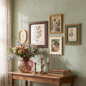 May include: A pink ribbed glass vase with a floral arrangement, four brass candlesticks, and a stack of three books. On the wall are five framed pictures, including botanical prints and portraits, featuring gold, dark brown, and green frames.
