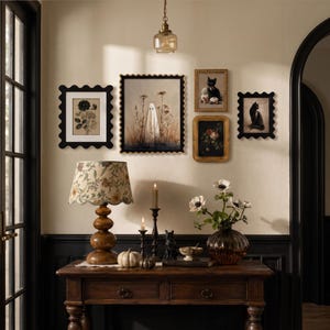 Op de afbeelding: Een houten sidetable bevat een lamp met een bloemenkap, een glazen vaas met witte bloemen, twee kaarsen en kleine pompoenen. Daarboven hangt een galerijmuur met vijf ingelijste kunstwerken, waaronder een geest en zwarte katten.