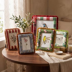 May include: A collection of five rectangular picture frames with fabric-covered borders and fringe trim. The frames feature various patterns including red stripes, blue floral, green floral with birds, and a red and white checkered design.