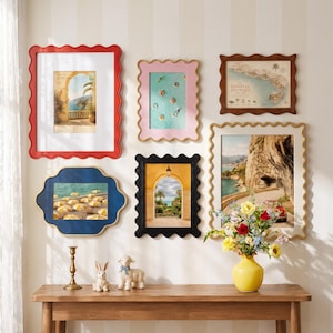 May include: A gallery wall arrangement featuring five scalloped picture frames in red, pink, dark blue, black, and cream, displaying coastal art prints. Below, a wooden console table holds a brass candlestick, two rabbit figurines, and a yellow vase.