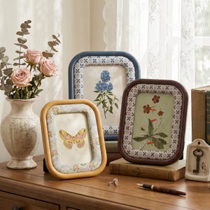 Puede incluir: Tres marcos de fotos rectangulares redondeados en amarillo, azul y marrón, cada uno con un borde estampado. Los marcos muestran ilustraciones botánicas y de insectos, incluyendo una mariposa amarilla, flores azules y un tallo floral rojo.