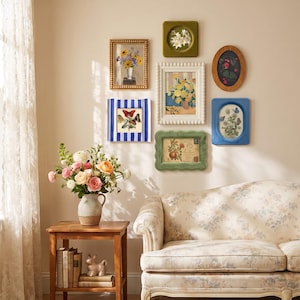 May include: A gallery wall featuring six framed art prints including floral and butterfly illustrations, arranged above a floral patterned sofa. A wooden side table holds a ceramic vase with fresh flowers and a stack of books.