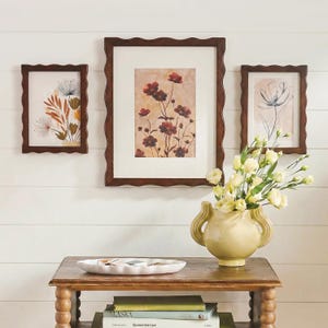 Könnte beinhalten: Drei dunkelbraune Rahmen mit gewelltem Rand und Blumenmotiven hängen über einem Holztisch. Eine hellgelbe Keramikvase mit zwei Henkeln enthält weiße Blumen. Ein weißes, gewelltes Tablett und gestapelte Bücher stehen auf der Tischoberfläche.
