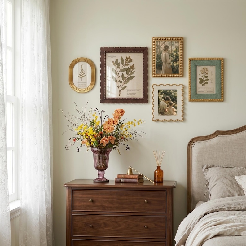 May include: A wooden dresser holds a purple glass vase filled with orange and yellow flowers, two stacked books with a small brass bell, and a glass reed diffuser. The wall features four framed botanical and portrait prints in gold and brown.