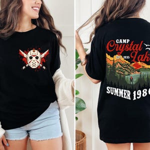 May include: Black t-shirt featuring a graphic of a hockey mask with crossed machetes and blood splatter. The back of the shirt displays a scenic design with the text "Camp Crystal Lake Summer 1980".