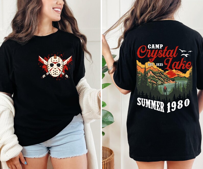 May include: Black t-shirt featuring a graphic of a hockey mask with crossed machetes and blood splatter. The back of the shirt displays a scenic design with the text "Camp Crystal Lake Summer 1980".