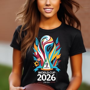 May include: Black t-shirt featuring a colorful graphic of a trophy, soccer ball, and the text "WORLD CUP 2026". The design includes vibrant hues of blue, orange, red, and yellow. The shirt is a short-sleeved crew neck.