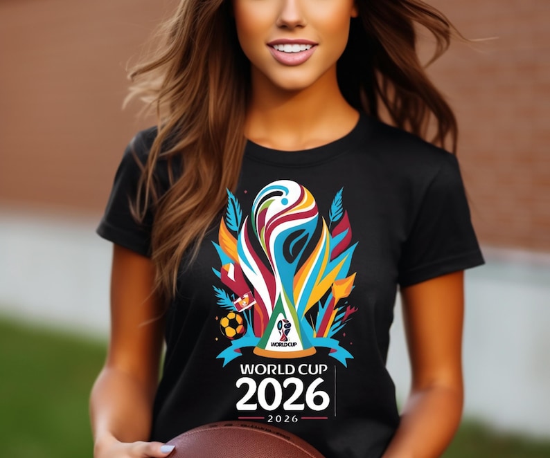 May include: Black t-shirt featuring a colorful graphic of a trophy, soccer ball, and the text "WORLD CUP 2026". The design includes vibrant hues of blue, orange, red, and yellow. The shirt is a short-sleeved crew neck.