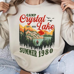 May include: Cream-colored sweatshirt with a graphic of a lake scene. The design includes the text "Camp Crystal Lake" in red, with "Est. 1935" and "Summer 1980" in green. The graphic features a lake, mountains, trees, and a person.