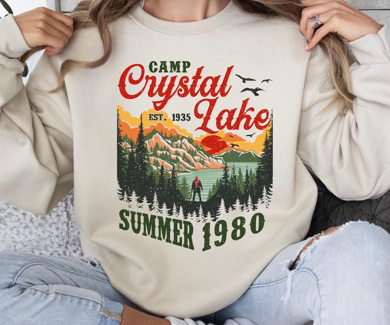 May include: Cream-colored sweatshirt with a graphic of a lake scene. The design includes the text "Camp Crystal Lake" in red, with "Est. 1935" and "Summer 1980" in green. The graphic features a lake, mountains, trees, and a person.