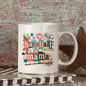 May include: White ceramic mug with the words "baseball mama" in a collage style design. The design includes a baseball, lips, stars, a megaphone, and a baseball bat. The mug is sitting on a notebook.