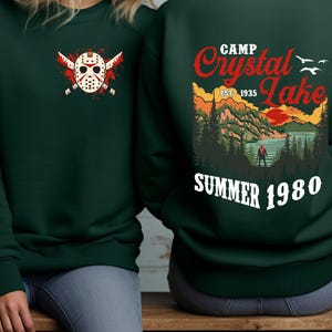 May include: Dark green sweatshirt with a graphic on the front of a hockey mask and crossed machetes. The back features a Camp Crystal Lake design with text that reads "Summer 1980".