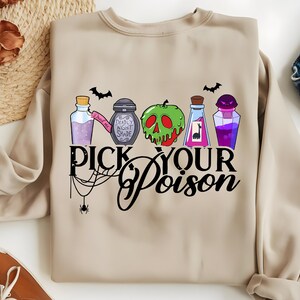 May include: Beige sweatshirt featuring a colorful graphic of potion bottles, a green apple with a skull design, and bats. The text "Pick Your Poison" is displayed in a stylized font, with a spider web and spider accent. A Halloween-themed design.