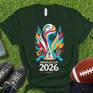 May include: Dark green t-shirt featuring a colorful graphic of the World Cup trophy with the text "WORLD CUP 2026". The shirt is on a green surface with a football and a pair of black shoes.