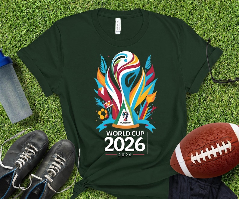 May include: Dark green t-shirt featuring a colorful graphic of the World Cup trophy with the text "WORLD CUP 2026". The shirt is on a green surface with a football and a pair of black shoes.