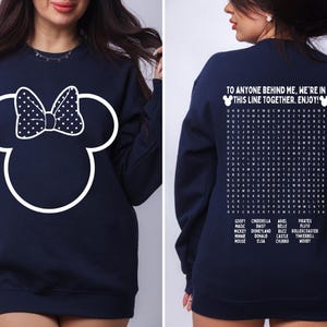 May include: Navy blue sweatshirt featuring a white Minnie Mouse outline with a polka dot bow on the front. The back of the sweatshirt has the text "TO ANYONE BEHIND ME, WE'RE IN THIS LINE TOGETHER. ENJOY!" and a word search.