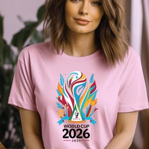 May include: A light pink t-shirt featuring a colorful graphic of a trophy with the text "WORLD CUP 2026". The graphic includes blue, red, yellow, and white elements. The shirt has short sleeves.