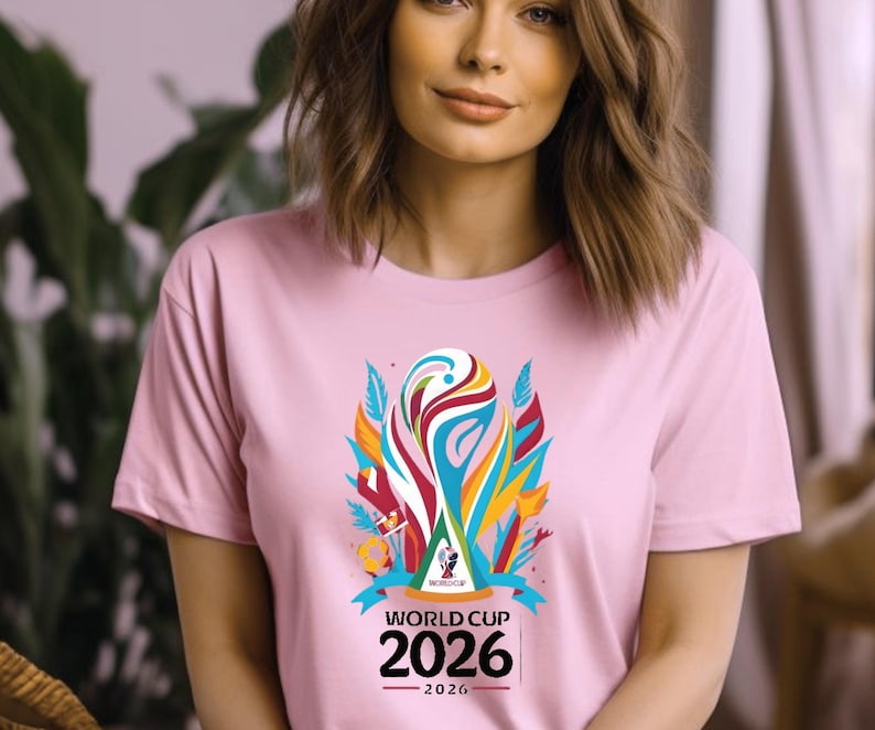 May include: A light pink t-shirt featuring a colorful graphic of a trophy with the text "WORLD CUP 2026". The graphic includes blue, red, yellow, and white elements. The shirt has short sleeves.