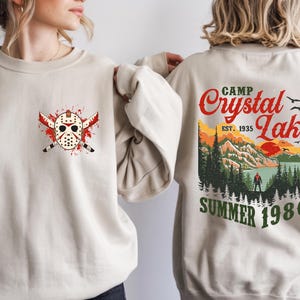 May include: Beige sweatshirt featuring a graphic of a hockey mask with crossed machetes and blood splatter. The back of the sweatshirt displays a scenic design with the text "Camp Crystal Lake Summer 1980".