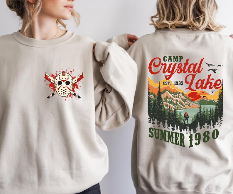 May include: Beige sweatshirt featuring a graphic of a hockey mask with crossed machetes and blood splatter. The back of the sweatshirt displays a scenic design with the text "Camp Crystal Lake Summer 1980".