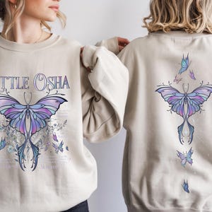 Peut inclure: Sweat-shirt beige avec un grand motif de papillon violet et bleu, et le texte "LITTLE OSHA". Le dos du sweat-shirt pr&eacute;sente &eacute;galement un motif de papillon. Le motif comprend des &eacute;l&eacute;ments floraux et de plus petits papillons.
