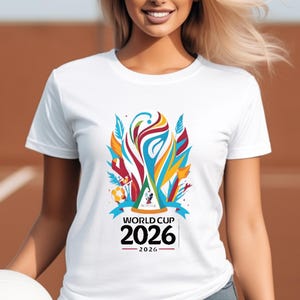 May include: White t-shirt featuring a colorful graphic design for the 2026 World Cup. The design includes a stylized trophy, ribbons, and the text "WORLD CUP 2026". The shirt is made of a soft material.