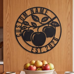 May include: A circular black metal wall sign featuring a cutout design of three apples on a branch. The text CUSTOM NAME is arched along the top left, and EST. 2026 is arched along the bottom right of the circular frame border.