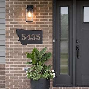 May include: A black metal house number sign shaped like the state of Montana, featuring the cutout numbers 5435. The sign is mounted on a tan brick wall next to a dark gray front door, a black outdoor light fixture, and a large potted green plant.