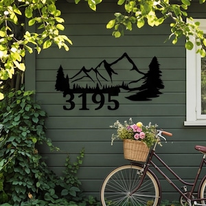 May include: A black metal wall sign featuring a mountain range silhouette with pine trees and the house number 3195. The sign is mounted on a dark green horizontal siding wall above a bicycle with a wicker basket filled with flowers.