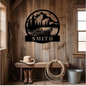 May include: A circular black metal wall sign featuring a laser-cut silhouette of a mountain range, pine trees, and a rowboat on water. The name SMITH is displayed in bold, capitalized letters on a rectangular banner at the bottom of the sign.