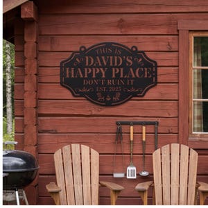 May include: A black metal wall sign with a decorative border featuring the text THIS IS DAVID'S HAPPY PLACE DON'T RUIN IT EST. 2025, accented with small chair and fire icons. The sign is mounted on a horizontal wooden plank wall surface.