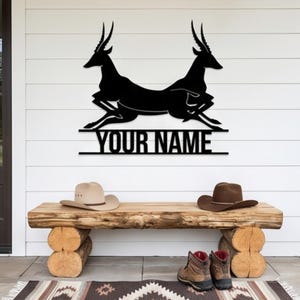 Pode incluir: Arte de parede em metal preto com duas gazelas correndo acima do texto "YOUR NAME". Abaixo da arte, há um banco de madeira com dois chapéus de cowboy e um par de botas. O fundo é uma parede branca.