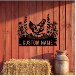 May include: A black metal wall sign featuring a silhouette of a chicken sitting on a nest of eggs, flanked by floral stems on both sides. Below the nest, a rectangular banner displays the text CUSTOM NAME in bold, capitalized lettering.