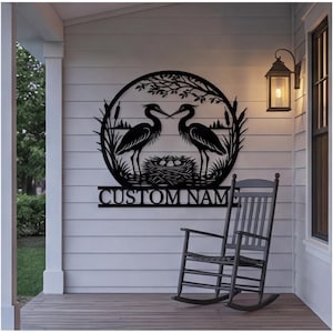 May include: A black metal wall sign featuring a circular frame with two herons standing by a nest of eggs, surrounded by reeds and tree branches. Below the circular design is a rectangular bar with the text CUSTOM NAME in capital letters.