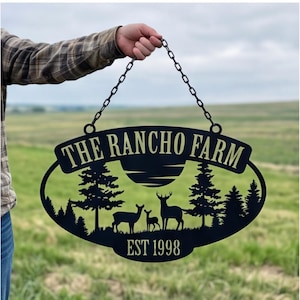 Puede incluir: Un letrero ovalado de metal negro con un diseño recortado de un bosque con tres ciervos y un sol. El texto dice THE RANCHO FARM y EST 1998. El letrero está suspendido por una cadena de metal negro, mostrando una silueta detallada de árboles.
