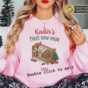 May include: A light pink sweatshirt with the text "Kadir's first new year" in red and green. A graphic of a baby in a crib with a Christmas tree and gifts is below the text. The year "1981" is printed below the graphic.
