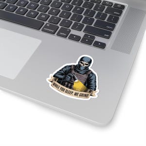May include: A sticker depicting a figure in tactical gear with a skull face, holding a flashlight. The figure is wearing a black balaclava and vest. The banner below reads "WHILE YOU SLEEP, WE GRIND."