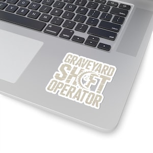 May include: A white sticker with the text "GRAVEYARD SHIFT OPERATOR" in a distressed, tan font. The sticker features a gear and a stylized face graphic. The sticker is placed on a silver laptop.