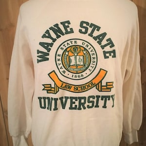 May include: Cream-colored Wayne State University sweatshirt with green and orange lettering. The design includes the university name, a circular emblem, and "Law School" text. The sweatshirt has long sleeves and ribbed cuffs.